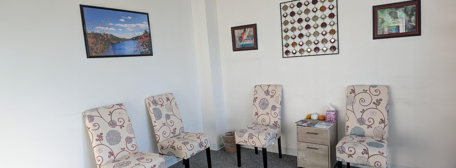 The image shows an interior space with a wall-mounted frame displaying multiple pictures, including a landscape scene and a close-up of a mug. Below the frame, there are two chairs facing each other, suggesting a seating area designed for conversation or interaction.